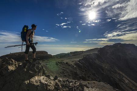 man with backpack standing on a cliff in mountains with blue sky and sunの写真素材