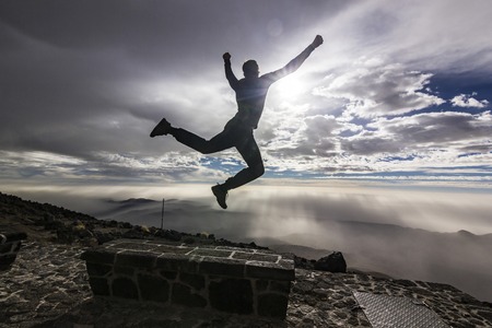 man jumping with hands up opposite sun with clouds on skyの写真素材