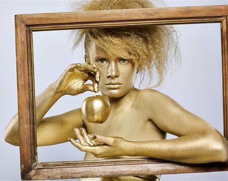 portrait of girl with golden bodyart posing with golden apple in her hands in picture frame on grayの写真素材