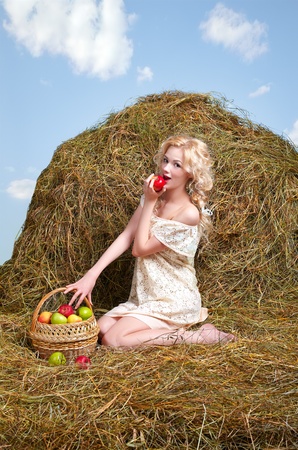 portrait of beautiful blonde country girl posing on yellow hay with basket of applesの写真素材