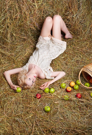 portrait of beautiful blonde country girl posing laying on yellow hay with apples spilled from basketの写真素材