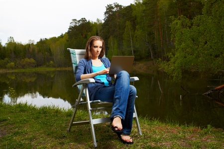outdoor portrait of beautiful woman sitting in folding chair with laptop near the pond in forestの写真素材