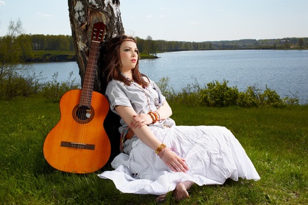 outdoor portrait of beautiful hippie girl sitting on green grass near birch with guitar. lake and forest on backgroundの写真素材