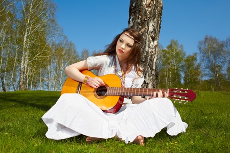 outdoor portrait of beautiful hippie girl sitting near birch on green grass with guitarの写真素材