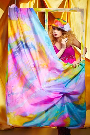 portrait of beautiful blonde woman artist in colorful hat standing behind the easel with painted cloth on itの写真素材