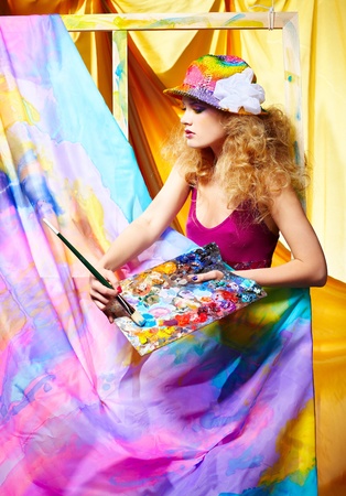 portrait of beautiful woman artist with paintbrush and palette standing behind the easel with painted cloth on itの写真素材