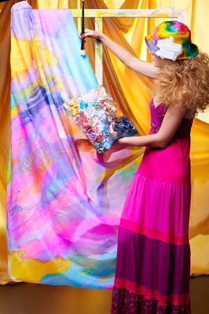 portrait of beautiful woman artist with paintbrush and palette standing in front of the easel with painted cloth on itの写真素材