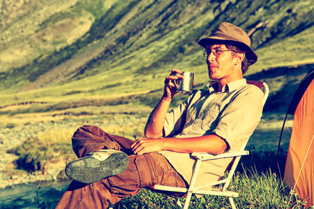 Colorized vintage outdoor portrait of man whith a pot in his hand is sitting in the chair at the Altai camp in mountains near the streamの写真素材