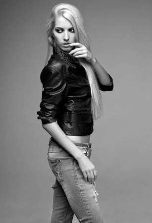 Monochrome portrait of blonde young woman in ragged jeans and black jacket on gray backgroundの写真素材