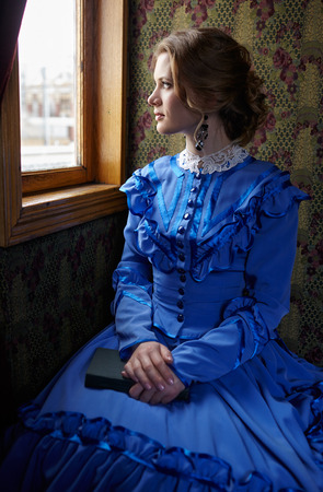 Young woman in blue vintage dress late 19th century sitting with book in coupe of retro railway train and looking out the windowの写真素材