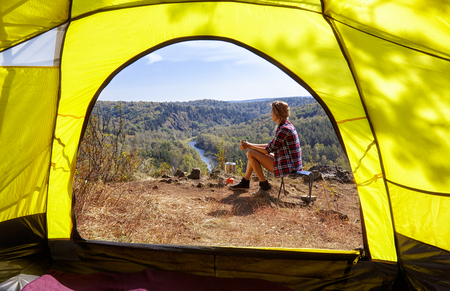 Young blonde woman tourists in camp on cliff over river and forest landscape. View from camping tent entrance.の写真素材