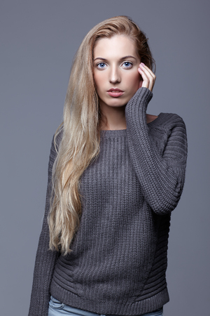 Portrait of young woman in gray woolen sweater and jeans. Beautiful girl posing on grey studio background. Female with blonde hair and day beauty makeup.の写真素材