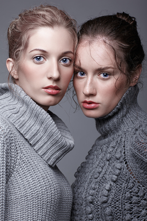 Two young women in gray sweaters on grey studio background. Beautiful girls stretching hands forward in embrace. Female friendship concept.の写真素材