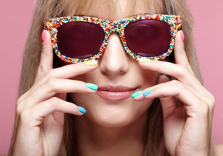 Young blonde smiling  woman with fun candy glasses and varicoloured finger nails manicure on pink background. の写真素材