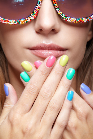 Young blonde woman with fun candy glasses and varicoloured finger nails manicure on pink background. の写真素材