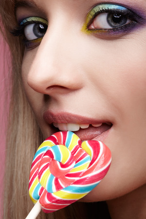 Beauty portrait of young blonde woman on pink background. Female with candy lollipop on stick in hands. Girl has finger nails with bright yellow, green, blue and pink manicure.の写真素材