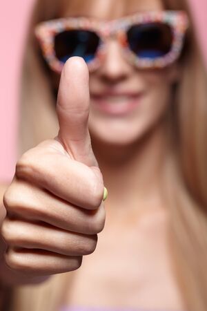 Female portrait with shallow depth of focus. Young blonde smiling  woman with fun candy glasses and thumb up on pink background. の写真素材