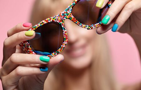 Female portrait with shallow depth of focus. Young blonde smiling  woman with huge fun candy glasses in hands on pink background. の写真素材