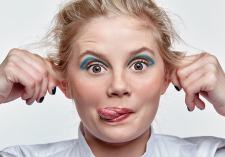 Emotional portrait of a young blonde woman on gray background. Female shows tongue and bulging her...