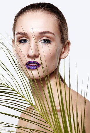 Female portrait with palm branch leaves on foreground. Woman has long neck and beauty face makeup with violet lips. Girl looking in camera with languid loving look.の写真素材