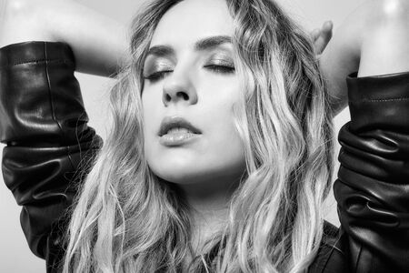 Monochrome portrait of young female with black leather jacket with eyes closed. Young pretty woman on gray background with hand behind headの写真素材
