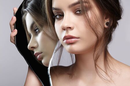 Girl with a shard of the mirror. Female with mirror shard in hand posing on gray background. Face reflection in mirror splinter.の写真素材