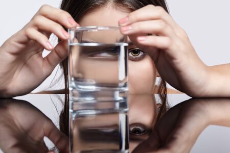 Girl hides her face behind a glass with water. Optical distortion  portrait of young woman at the mirror table. Female on gray background.の写真素材