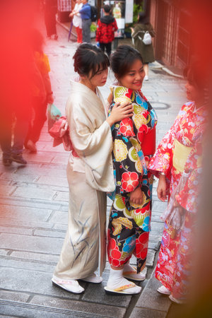 KYOTO, JAPAN - OCTOBER 18, 2019: Young girls wearing kimono on the street of old Kyoto neighborhood. The view through the blur autumn leaves with front bokeh effect. Kyoto, Japan.のeditorial素材