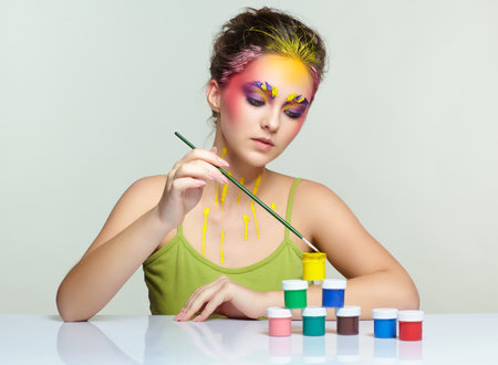 Portrait of young woman posing at the table with pyramid of small cans of paint and brush in hand. Unusual female art make-up with paint on brows, hair and around eyes.の写真素材