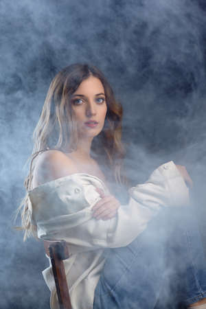 Portrait of young beautiful blonde woman is sitting on a chair. Female on smoke background.の写真素材