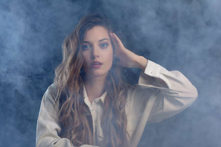 Portrait of young beautiful blonde woman is sitting on a chair. Female on smoke background.の写真素材