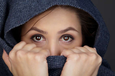 Beauty portrait of brunette woman dressed in dark blue scarf. Female portrait from a three-quarter angle on black background. Hands cover face with a scarfの写真素材