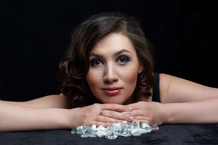 Portrait of beautiful long-haired brunette woman on black background. Female sit at table covered by black velvet. There are glass stones on the table.の写真素材