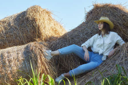 Young brunette woman in a white shirt and jeans is sitting on the rolls of hay. Romantic country girl in cowboy hat on hay.の写真素材