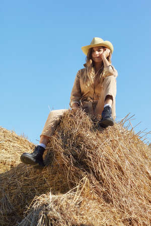 Young blonde woman in a beige jumpsuit is sitting on the rolls of hay. Romantic country girl in...