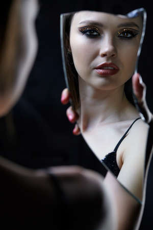Female looks into a shard of the mirror. Beautiful young woman portrait with vogue shining face makeup. Eye shadows with golden sparcles.の写真素材
