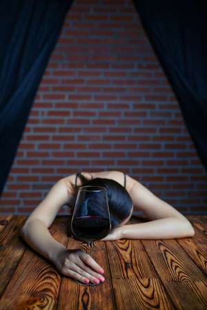 Portrait of a beautiful young brunette woman sleeping at the wooden table with a glass of red wine. In the background there is a brick wall and black curtains.の写真素材