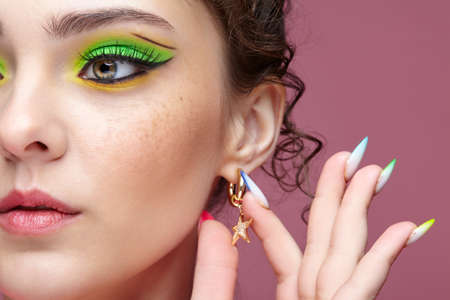 Portrait of a young woman touches an earring in the form of a star on her ear. Female with unusual green eyes makeup. Girl with nails with multi-colored manicure.の写真素材