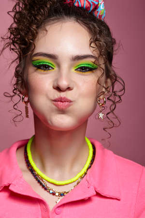 Girl puffs out her cheeks. Portrait of young woman dressed in pink dress on pink background. Female with unusual green eyes shadows makeup, curly hair and earrings.の写真素材