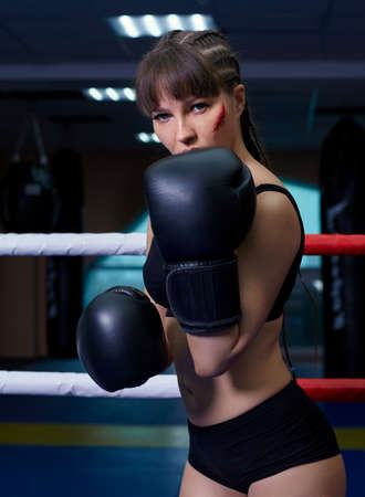 Female fighter with boxing gloves on her hands stands in a boxing ring. Woman has an abrasion on her cheek.の写真素材