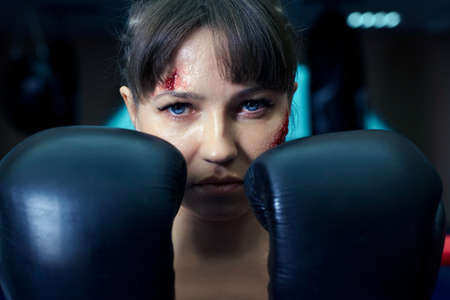 Female fighter with boxing gloves on her hands stands in a boxing ring. Woman has an abrasion on her cheek.の写真素材