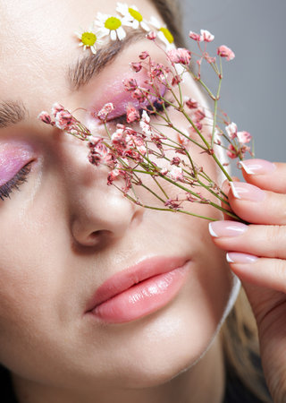 Close up portrait of female face with pink makeup. Woman with Gypsophila or babe's breath flowers in hand near face. Brows are decorated by small chamomile flowers. eyes closed.の写真素材