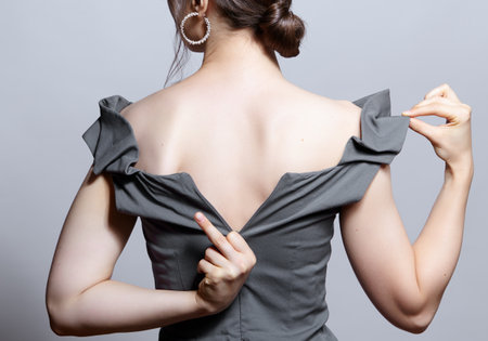 Young woman unzips the zipper on the dress. Hand showing fuck sign. view from the back. Bunette female rear view with hair knot and earrings in the ears.の写真素材