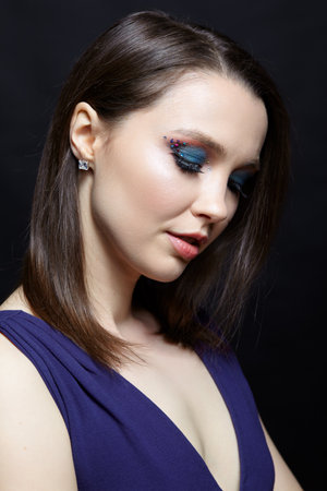 Portrait of beautiful brunette female with blue eye shadow make up and rhinestones. Young woman in blue dress on black background.の写真素材