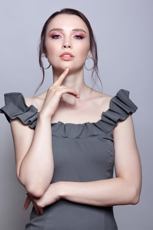 Portrait of young woman in gray dress on gray background. Bunette female with hands near face and earring in the ear. beauty makeup concept.の写真素材