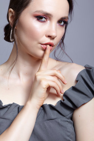 Portrait of brunette woman on gray background. Female in gray dress with pink smoky eyes eye shadows. Finger on lips with silence sign.の写真素材