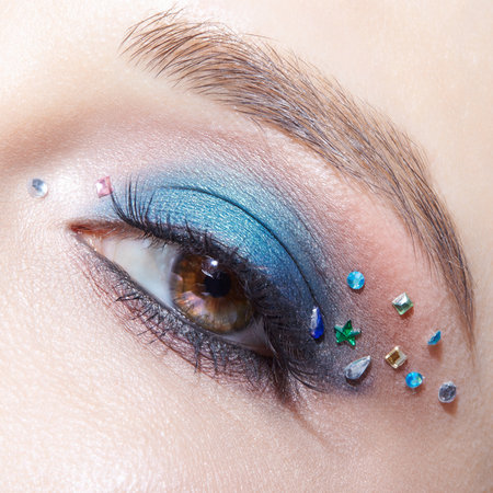 Closeup macro shot of human female brown color eye. Woman with natural evening vogue face beauty makeup. Girl with perfect skin, with blue eye shadow make up and rhinestones.の写真素材