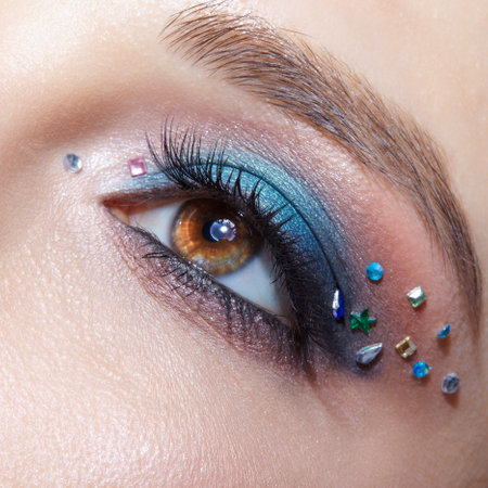 Closeup macro shot of human female brown color eye. Woman with natural evening vogue face beauty makeup. Girl with perfect skin, with blue eye shadow make up and rhinestones.の写真素材
