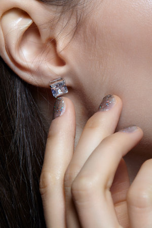 Portrait of a young woman on gray background. Female posing with hand near face. In the ear is an earring with a white crystal stone. Nails covered in glittering glitter manicure.の写真素材