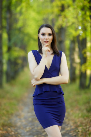 Female in blue dress walking along autumn linden tree alley. Path under yellow trees with falling autumn leaves. Sunny autumn day. The rays of the sun shine through the trunks and crowns of trees.の写真素材